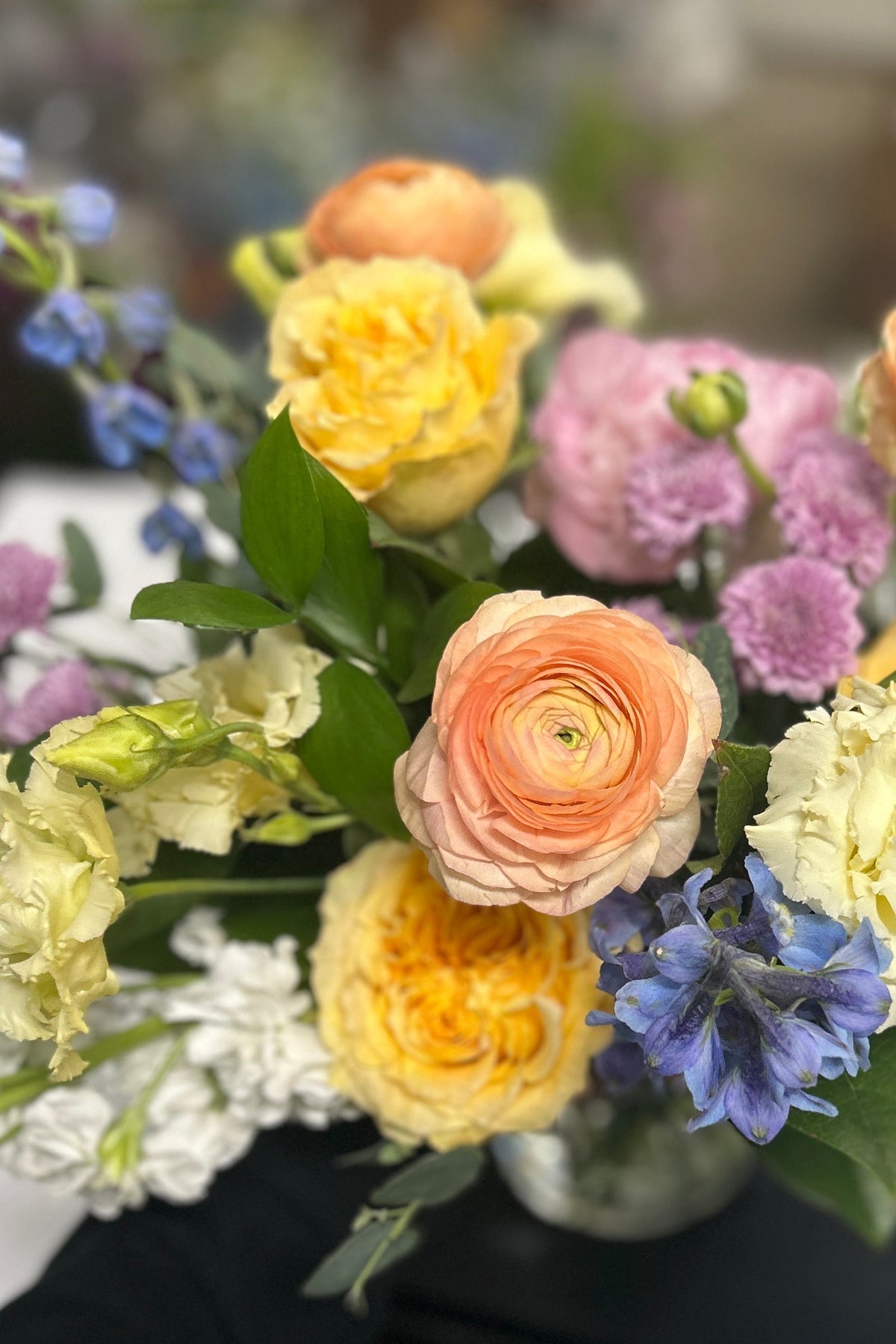 A colorful mixed flower bouquet held by a person wearing a black sleeve, featuring peach roses, yellow ranunculus, lavender carnations, blue delphiniums, and white baby's breath in a glass vase.
