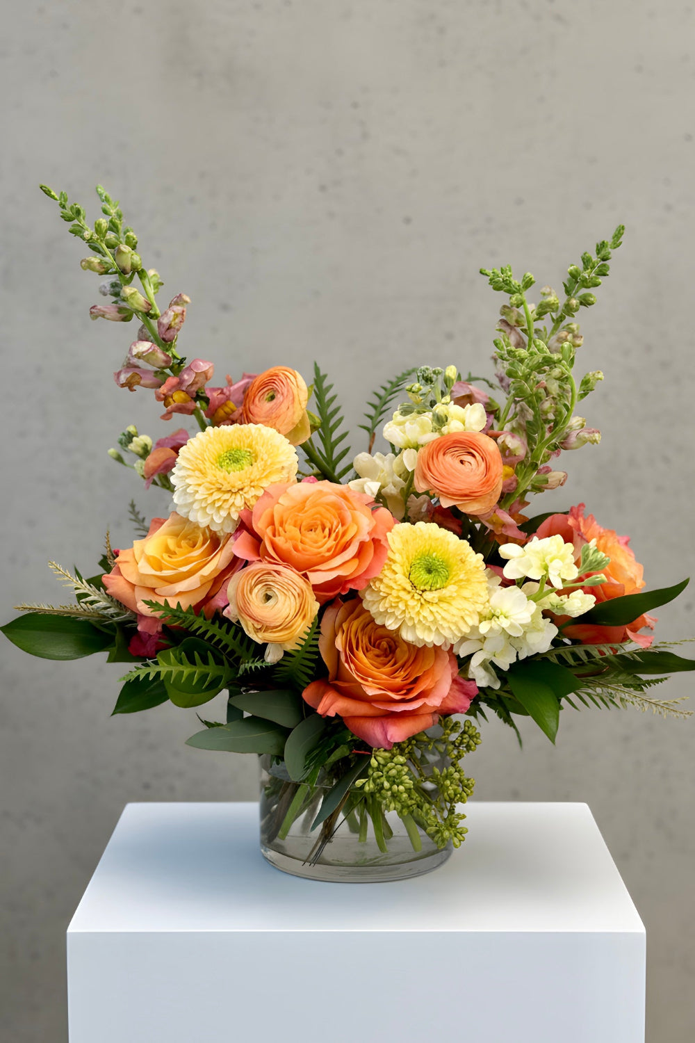A vibrant floral arrangement featuring peach roses, yellow ranunculus, and green foliage in a clear glass vase, displayed on a white pedestal against a textured gray background.