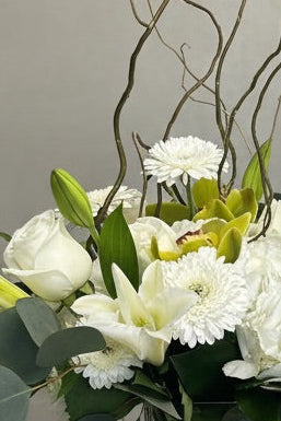A floral arrangement featuring white lilies, white daisies, and green foliage in a clear glass vase, with tall bare twigs rising above, displayed on a white pedestal against a textured gray wall.