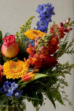 A vibrant autumn floral arrangement in a clear glass vase, featuring orange and red roses, yellow daisies, blue delphiniums, and green foliage, displayed on a white pedestal against a neutral concrete wall.