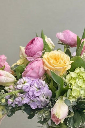 A vibrant flower bouquet in a clear glass vase, displayed on a white pedestal against a neutral gray background. The bouquet features a mix of pink, yellow, and purple flowers with green foliage.