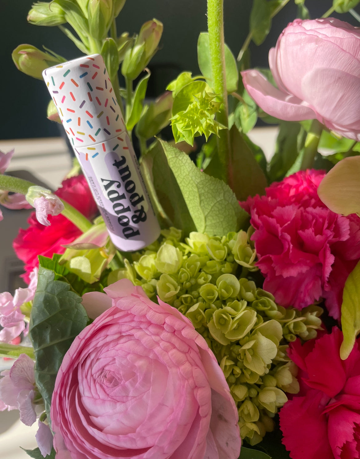 A custom flower arrangement with a 'Poppy & Pout' lip balm tub placed among the blumes. The arrangement is composed of pink and green flowers, with a purple and white striped lip balm tube.