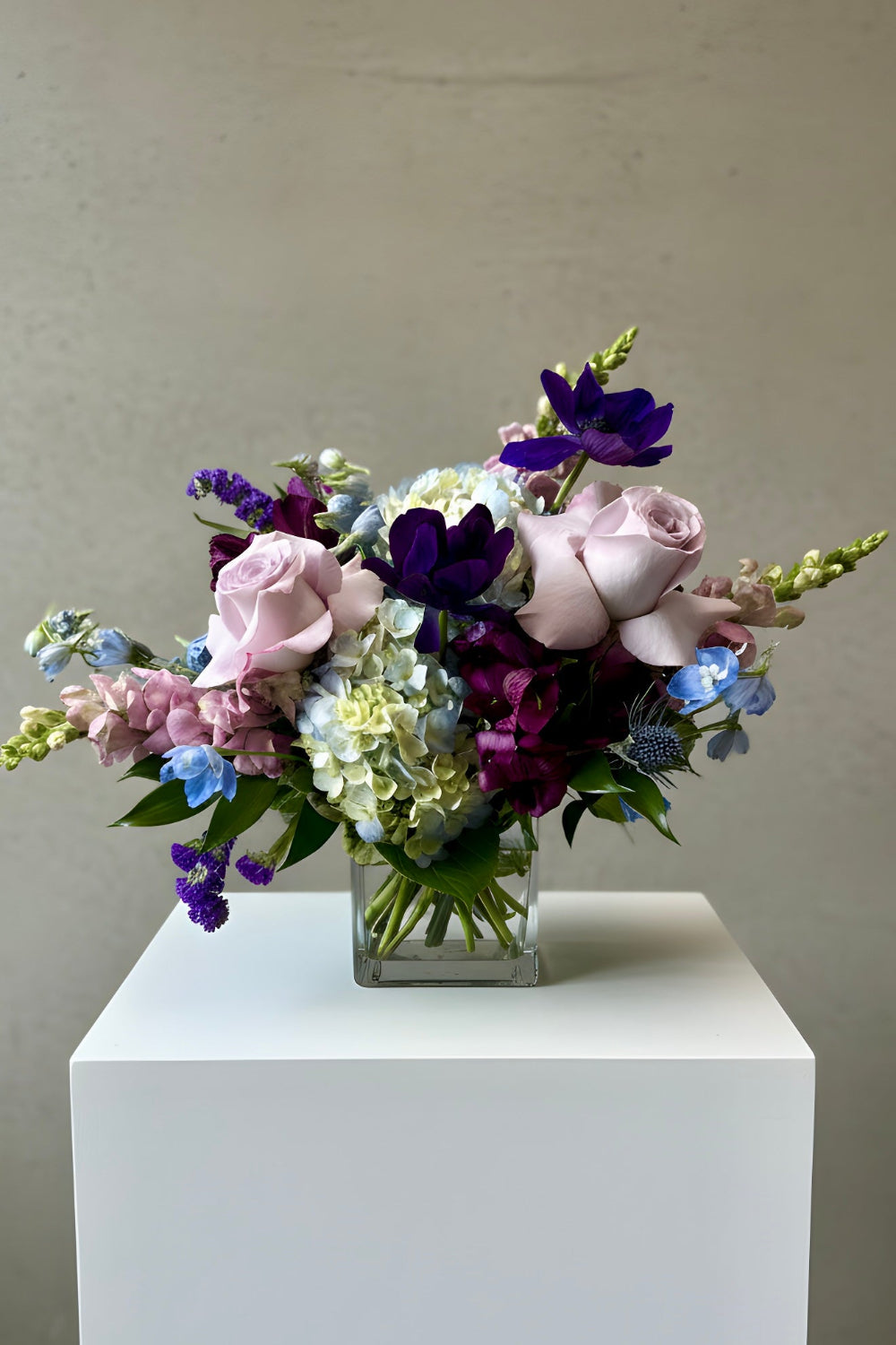 A floral arrangement of pink, purple, blue, and white flowers in a clear square glass vase, displayed on a white pedestal against a neutral beige wall.