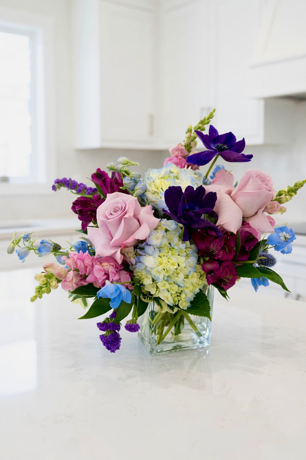 A vibrant floral arrangement featuring pink roses, deep purple anemones, light blue delphiniums, and white hydrangeas in a clear glass vase, set on a white marble kitchen counter.