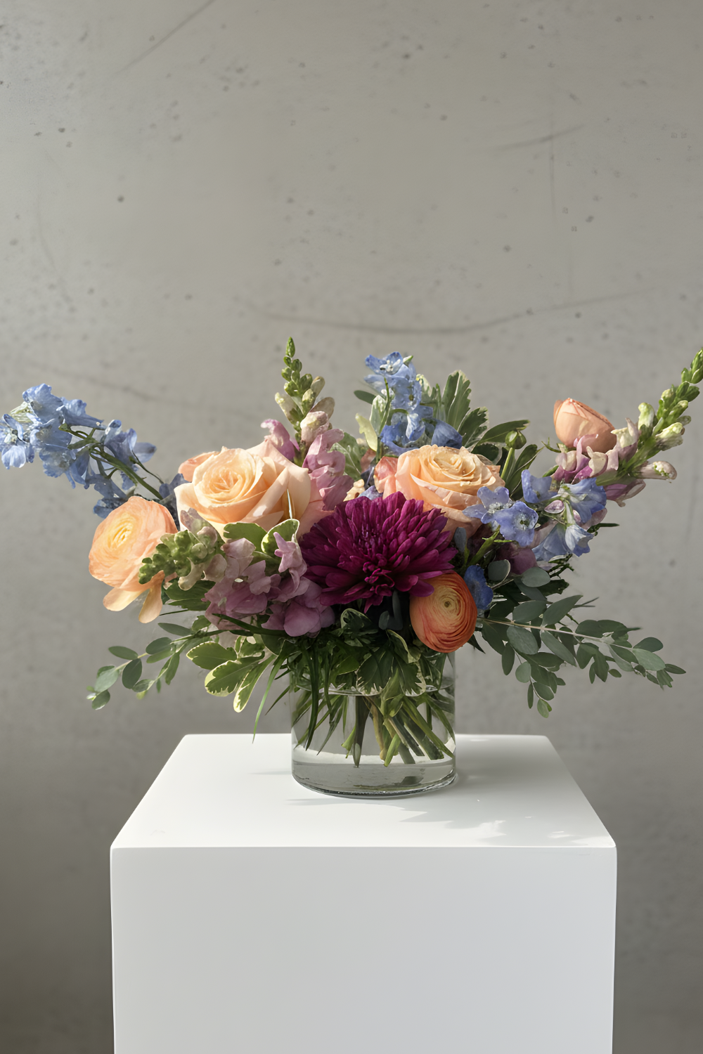 A vibrant mixed floral arrangement in a clear glass vase, displayed on a white pedestal against a textured gray concrete wall. The bouquet features peach roses, light blue delphiniums, a deep purple chrysanthemum, and green foliage.