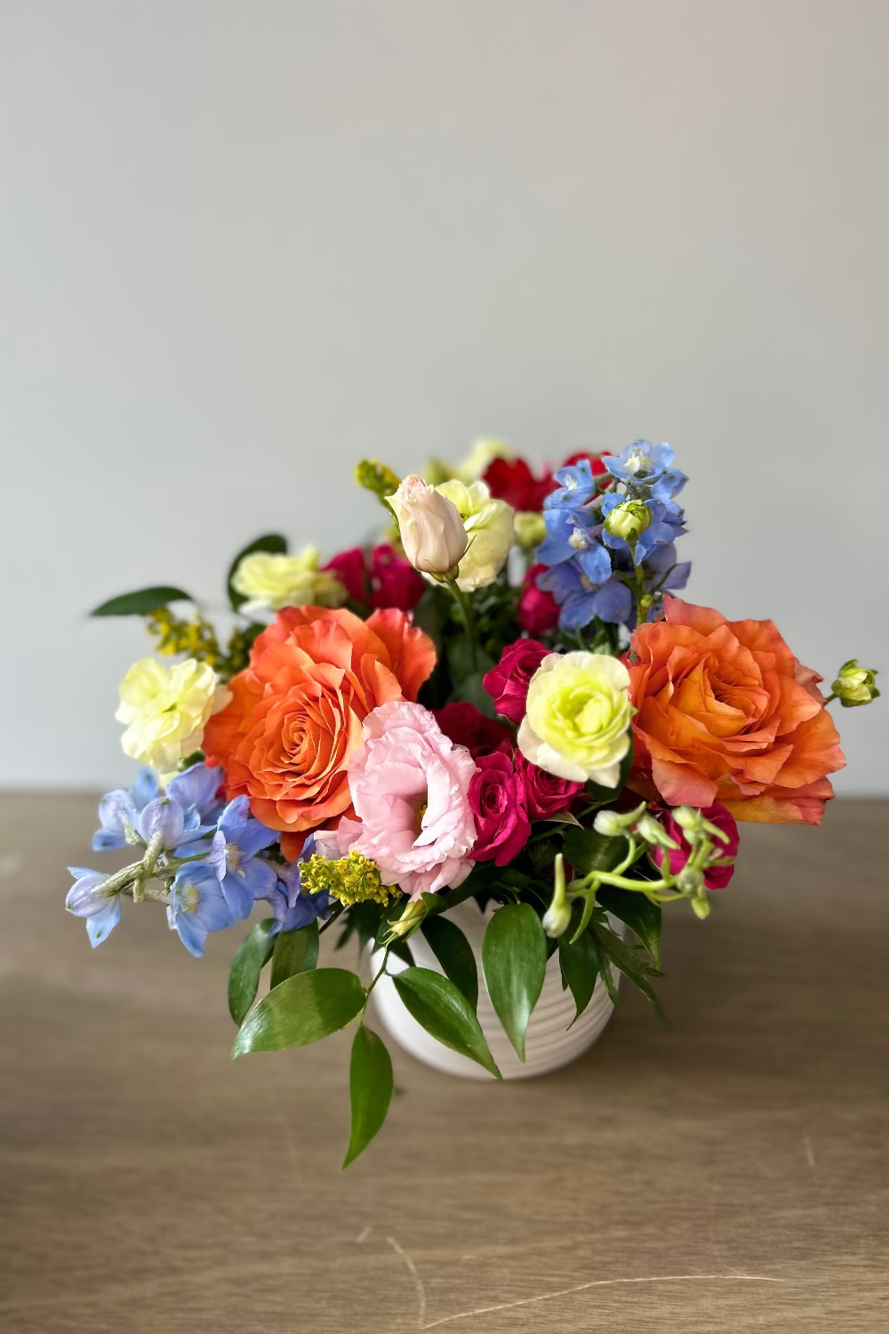 A vibrant mixed flower bouquet in a small white ceramic vase, featuring orange and pink roses, blue delphiniums, yellow lisianthus, and green foliage, set on a wooden tabletop against a neutral gray background.