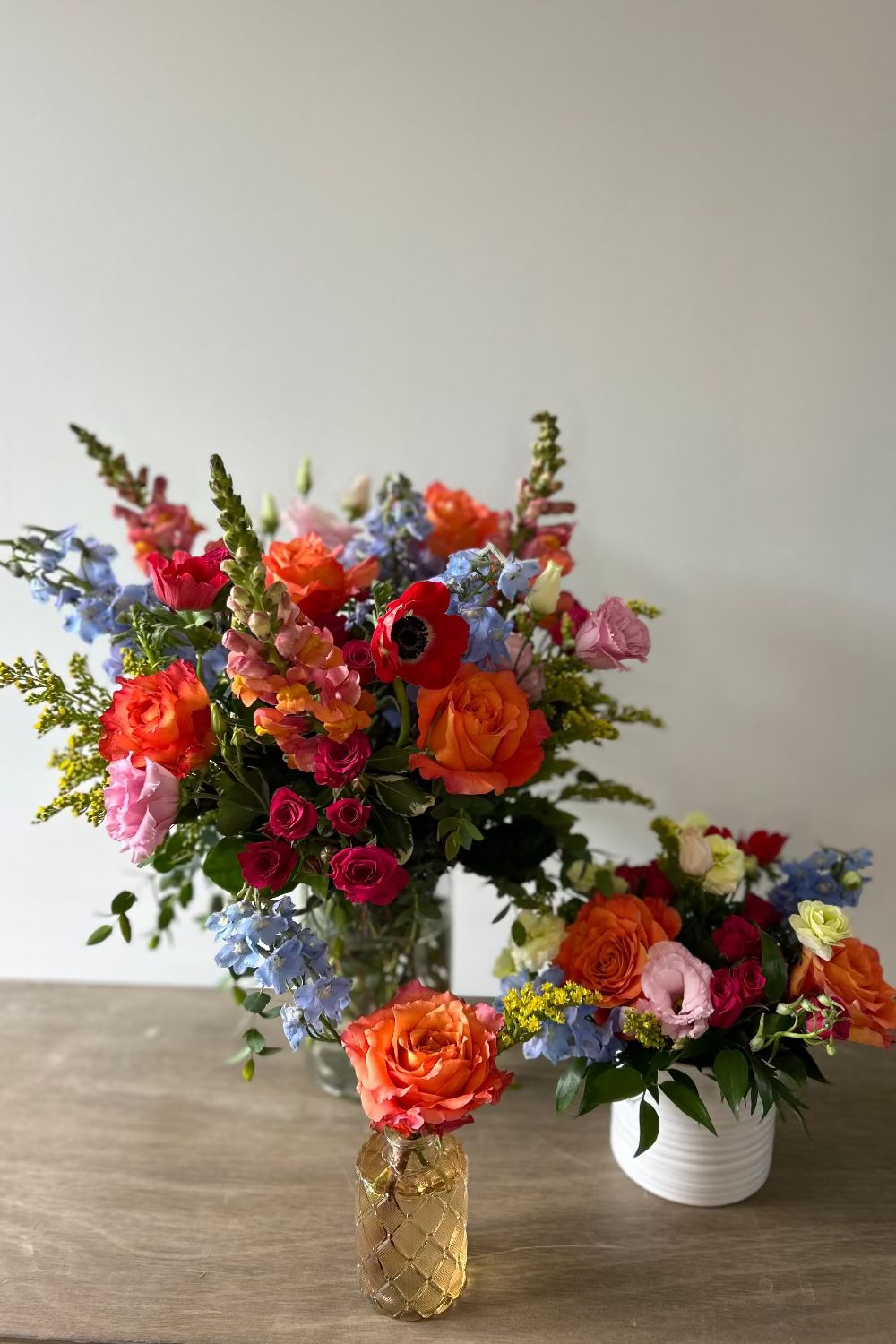 A vibrant floral arrangement featuring orange roses, pink and red flowers, blue delphiniums, and green foliage, styled in a glass vase on a wooden table next to a smaller matching arrangement in a white pot.