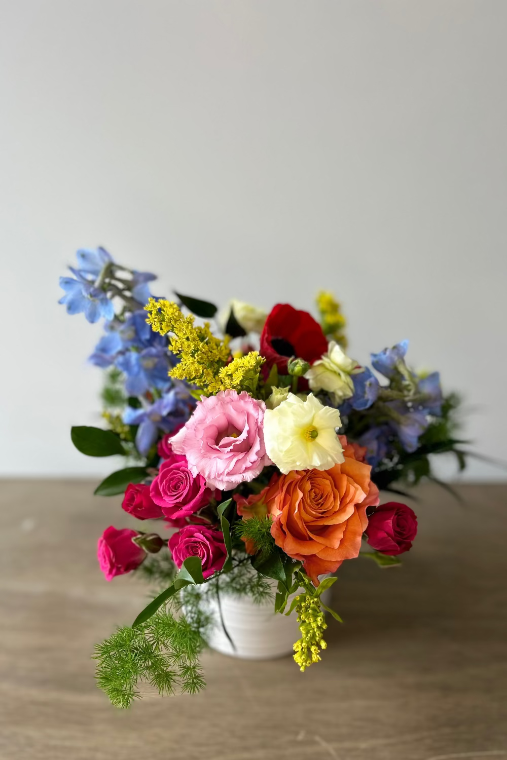 A vibrant mixed flower bouquet in a small white ceramic pot, featuring pink, orange, red, blue, yellow, and white flowers, set on a light brown wooden table against a neutral gray wall.