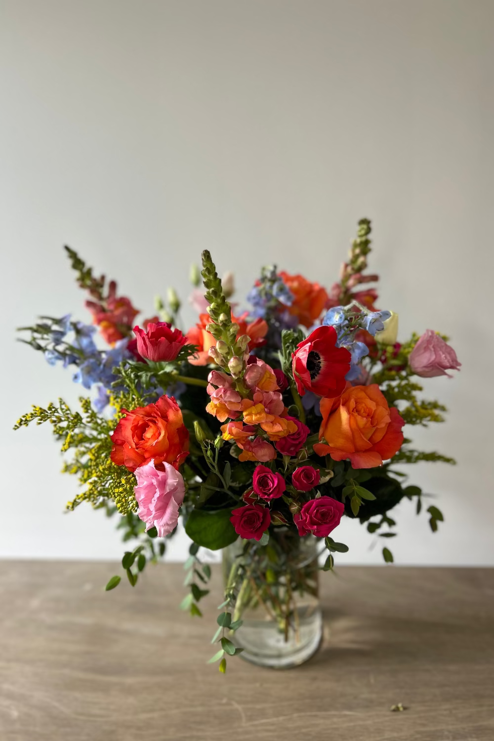 A vibrant mixed flower bouquet in a clear glass vase, featuring orange and pink roses, red poppies, blue delphiniums, and green foliage, set on a wooden tabletop against a neutral light gray background.