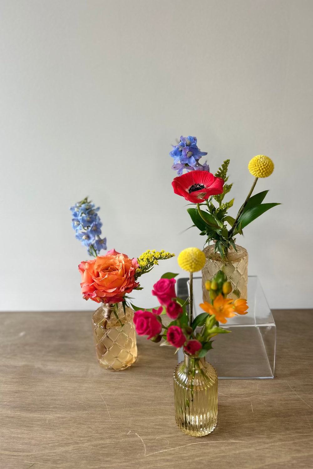 Three small clear glass vases with textured patterns holding colorful spring flowers, arranged on a wooden tabletop against a plain light gray wall.