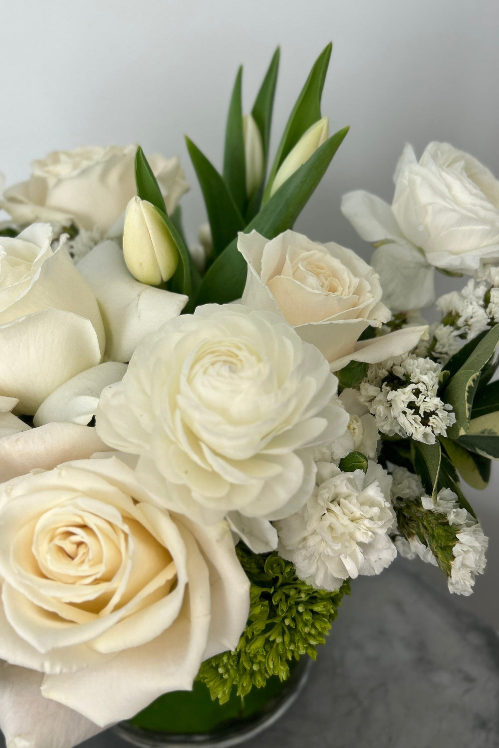 A bouquet of white and cream roses with green foliage and small white filler flowers, arranged in a clear vase on a gray surface against a light gray wall.