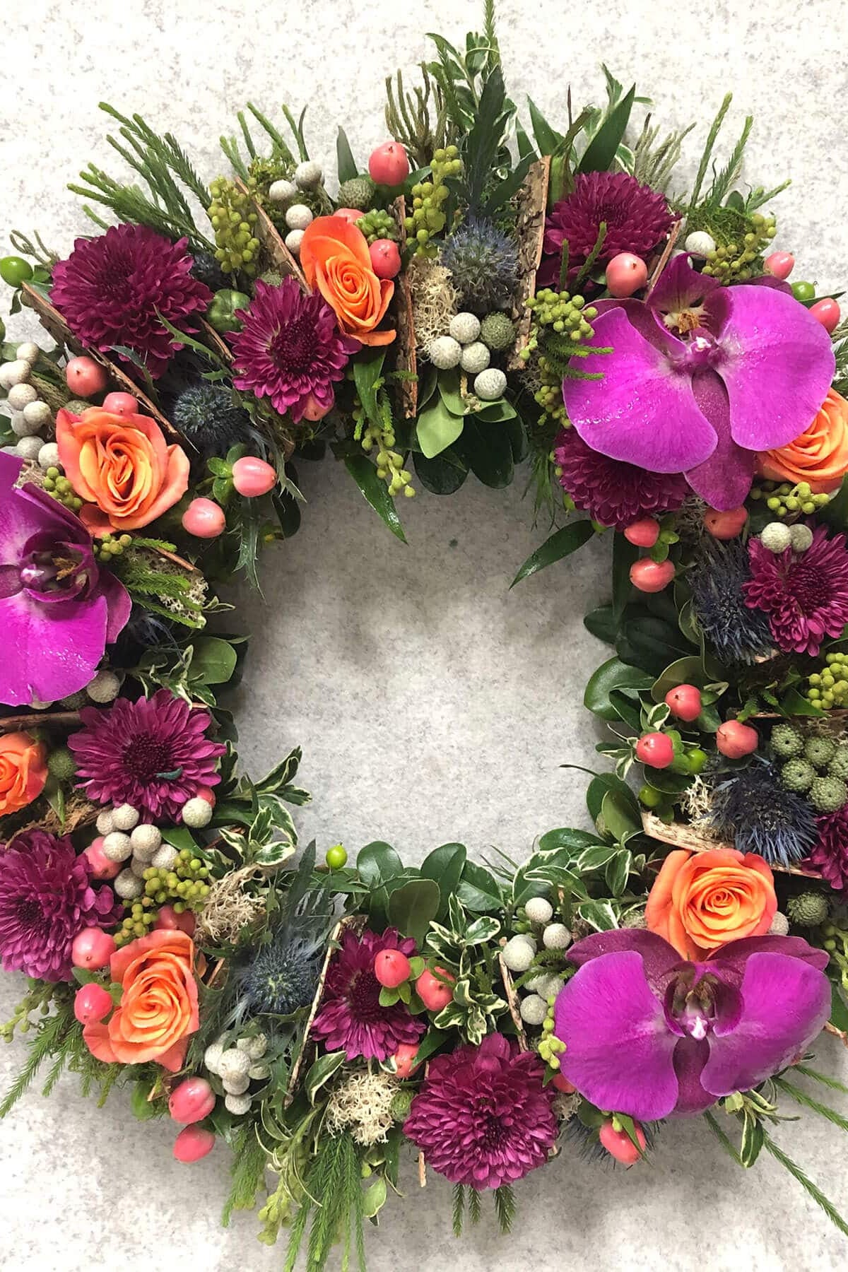 A round floral wreath with a mix of purple, pink, and orange flowers, green foliage, and natural elements.