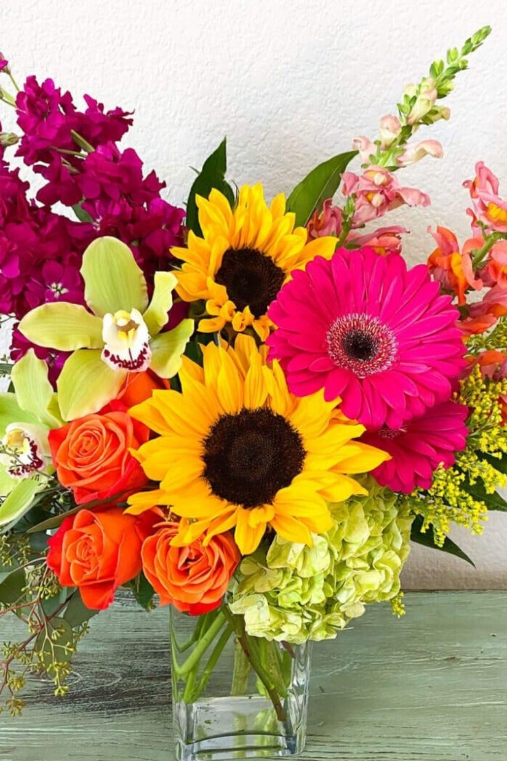 A colorful floral bouquet featuring sunflowers, roses, and orchids in a clear glass vase, designed to represent the Bright Blooms product.