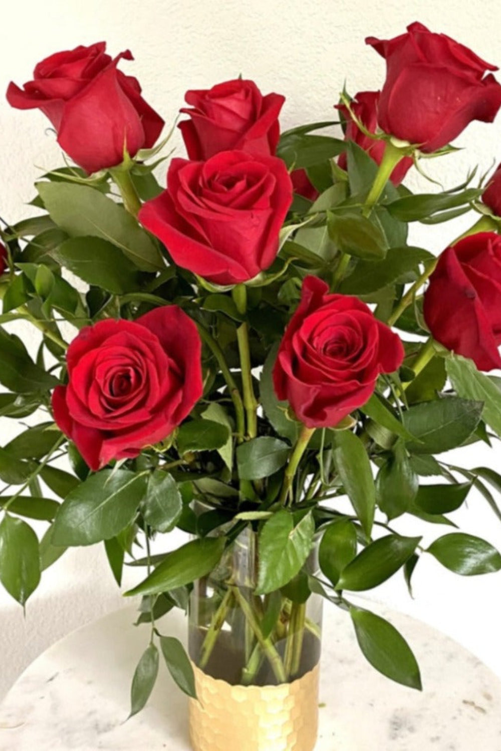 A dozen red roses arranged in a clear vase with green leaves.